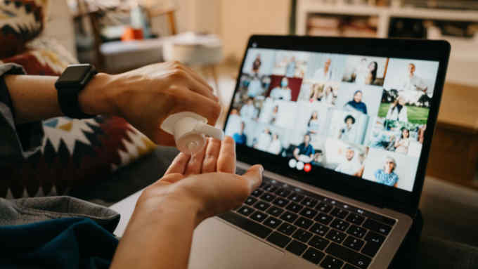 Woman is using hand sanitizer while she's having a video call with many people on her laptop. She's at home during coronavirus Covid-19 pandemic quarantine.