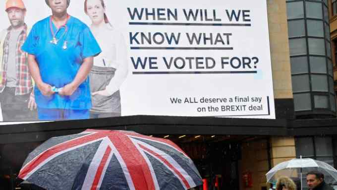 A pedestrian shields himself with a British union flag umbrella as he passes a billboard with a Brexit related campaign slogan in Leicester Square, London, Britain, March 29, 2018.  REUTERS/Toby Melville - RC1241766BF0