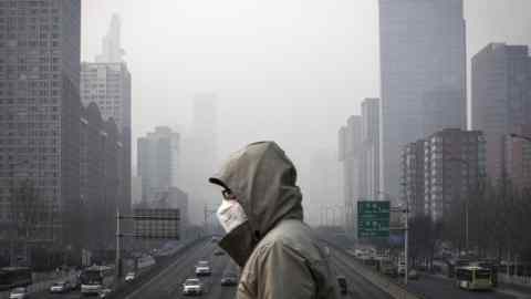 A man wearing a face mask walks on a footbridge as buildings shrouded in haze stands in the background in Beijing, China, on Friday, Jan. 6, 2017. Toxic haze that settled over much of China during the last three weeks has triggered a flight reflex among residents, leading to the rising popularity of smog avoidance travel packages to far-flung locations such as Iceland and Antarctica. Photographer: Qilai Shen/Bloomberg