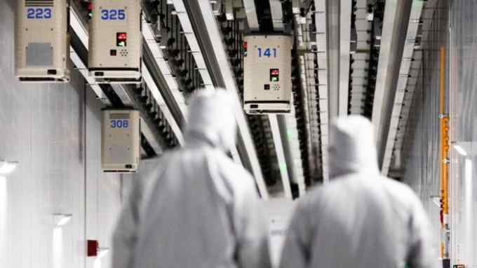 Employees wearing cleanroom suits walk beneath Automated Material Handling Systems (AMHS) vehicle robots moving along tracks on the ceiling inside the GlobalFoundries semiconductor manufacturing facility in Malta, New York