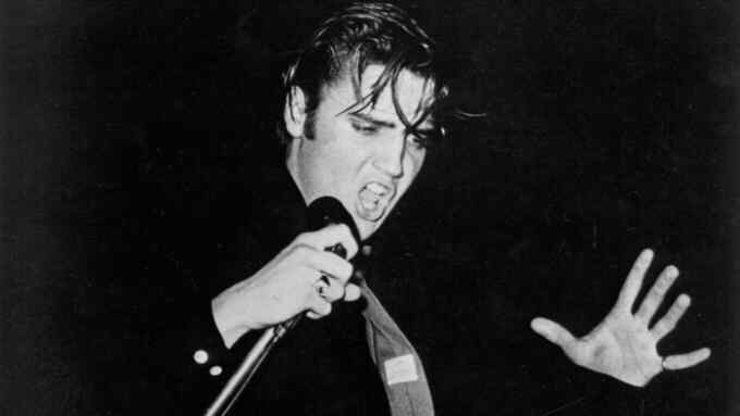 Elvis Presley on stage singing into a microphone, his hair flopping over his forehead, one hand extended