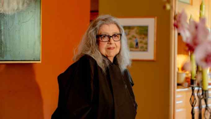 An older woman with long grey hair wearing a black, wide shirt and glasses stands in her living room against an orange background.