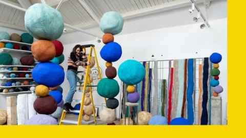 A large white room with tall sculptures of coloured balls stacked on each other. A woman stands on a ladder among them