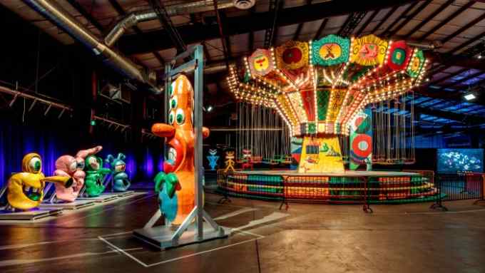 In a large warehosue, colourful, totemic monster sculptures surround an illuminatead merry-go-round