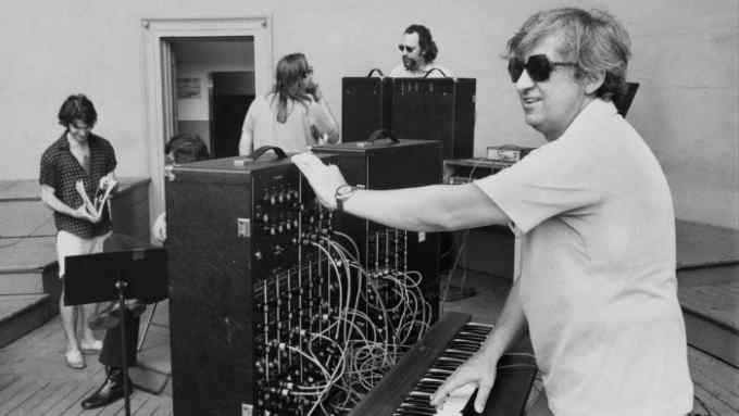 A man wearing dark glasses stands in a recording studio at the keyboard of a synthesiser which sprouts wires and plugs