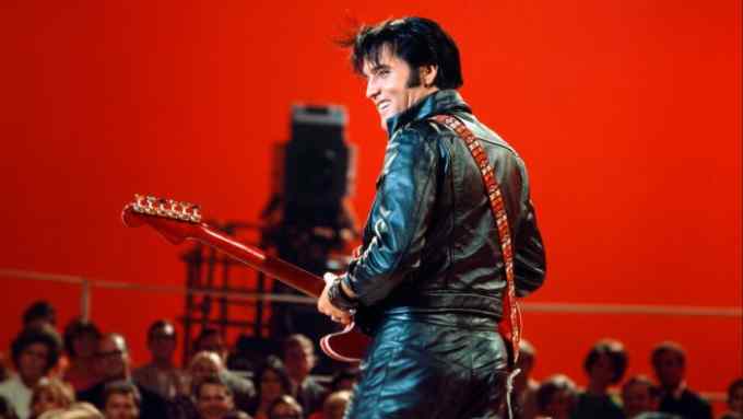 Seen from behind, Elvis Presley, wearing black leather and playing an electric guitar, stands smiling against a red background