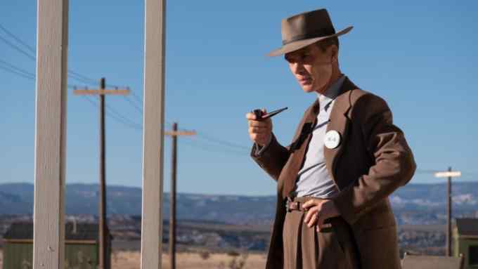 A man in a mid-brown 1950s suit and fedora stands holding a pipe in front of a desert landscape
