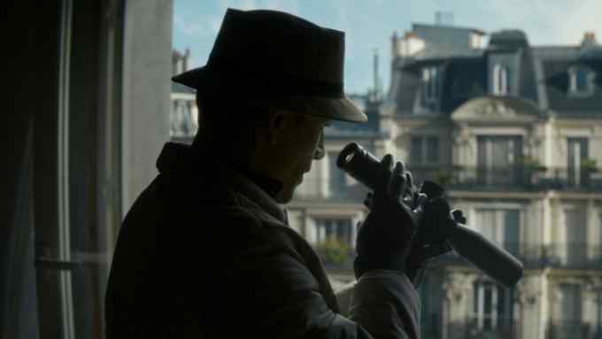 A man in a bucket hat and gloves look through a scope across a Paris street