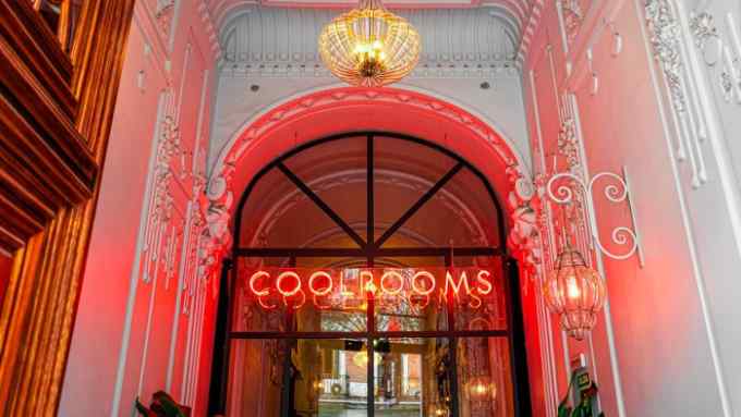 A red neon sign that reads ‘CoolRooms’ in a large arched window flanked by neoclassical cornicing