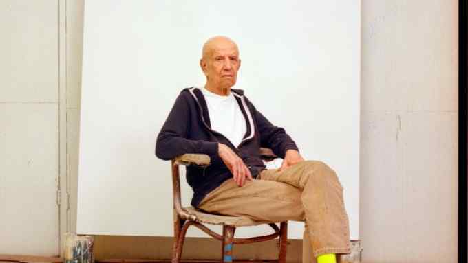 An elderly man wearing a blue zip-up hoodie, a white T-shirt and beige trousers sits on a chair in the centre of a room