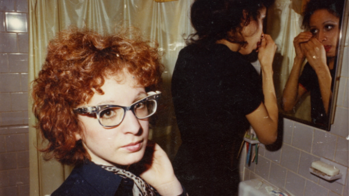 A pale woman with red hair and glasses looks at the camera as another woman looks into a bathroom mirror