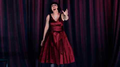 A woman in red dress stands singing in front of red curtains, one arm outstretched