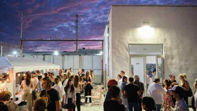 People drink and chat outside a white-walled gallery with a purple sky