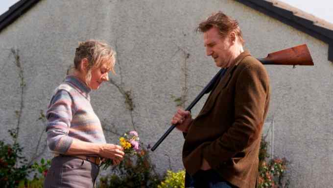 Next to the concrete wall of a building, a woman and a man stand facing each other; she looks down at a small bunch of wild flowers in her hands; he looks at her, and has a shotgun over his shoulder