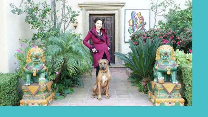 A woman in a smart cerise jacket standing behind a dog and in front of a large house