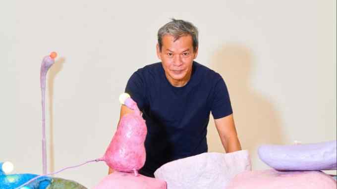 A man in a navy T-shirt stands behind a table covered in a dozen candy-coloured objects and vessels, some with light fixtures. He holds a pink object