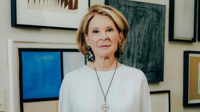A woman in a white top and black trousers stands in front of paintings
