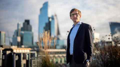 Chris Miller in suit and open-necked blue shirt standing on the roof of a building with an out-of-focus view of the City of London in the background