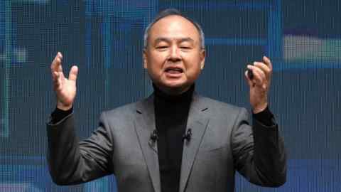 Masayoshi Son, wearing a gray suit and black turtleneck, speaks with his hands raised