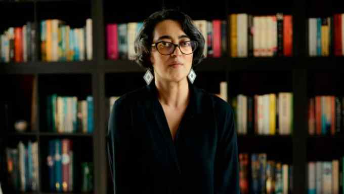 A woman with black glasses and medium-length dark hair in front of full bookshelves looks suspiciously at the camera