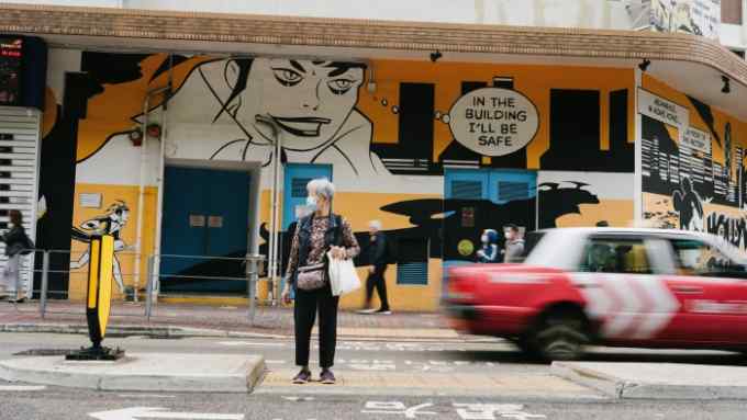 An older woman stands at a crossing as a taxi speeds behind her in front of a building covered in street art. In the art, a woman’s speech bubble says: ‘In the building I’ll be safe’