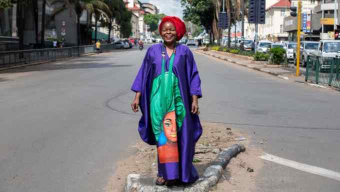 A woman in bright gown and headscarf stands in the middle of an empty road, smiling