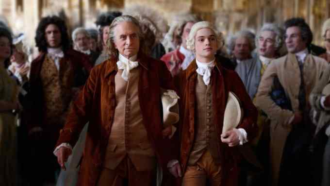 A distinguished-looking man with long grey hair wearing a cravat, rust-coloured jacket and waistcoat walks next to a teenage boy wearing similar clothing and a white wig. Behind them is a crowd of men and women dressed in opulent 18th-century attire.