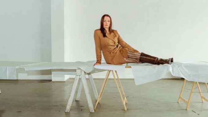 A young woman wearing a beige double-breasted blazer dress, chequered tights and brown boots sits on a long, wooden working desk in a white studio