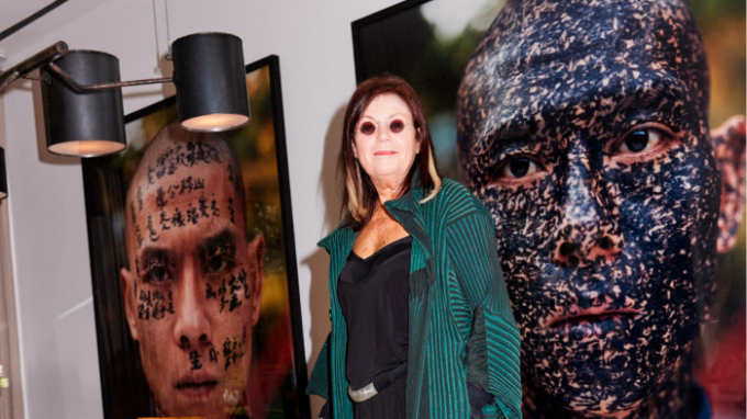 A woman wearing a pleated dark green jacket stands in front of a photo of a man whose face is covered in calligraphy