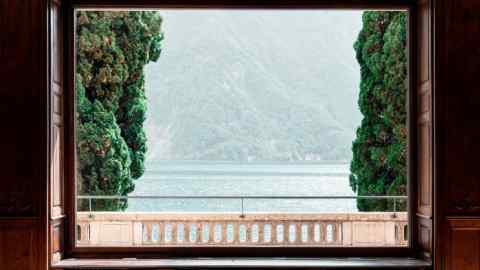A wide, lake-facing window offers a scenic view of a lush hilly landscape, enclosed by cypresses and shimmering water