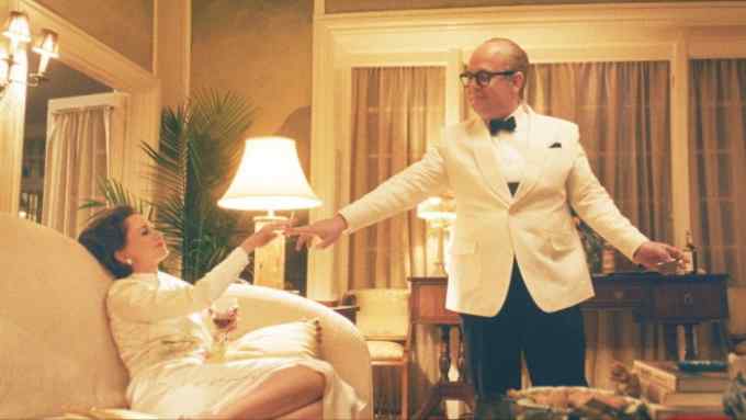 An elegant woman in a white dress sits with her feet up on a sofa with a glass of wine. She takes a cigarette from a man wearing glasses and a white tuxedo, who stands next to her in a stylish living room.