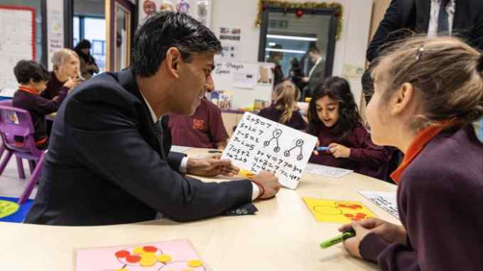Rishi Sunak sits at a little desk with a year 1 pupil looking at a maths problem on a small whiteboard