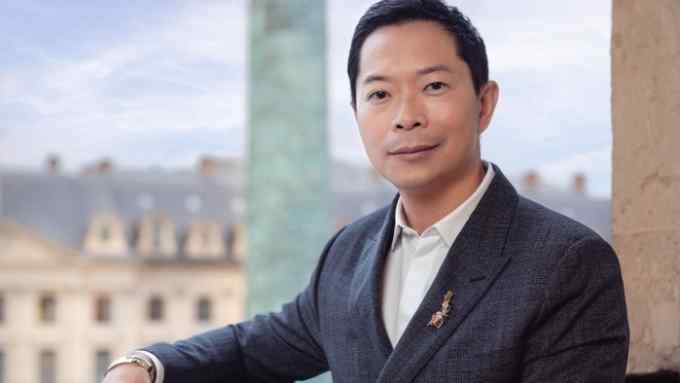 A portrait of a well-dressed man of Asian descent, leaning on a balcony railing with the Place Vendôme Column visible behind him