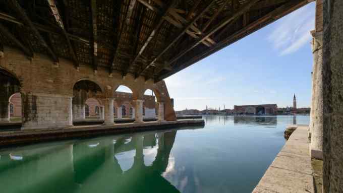 A photograph depicts the water-overlooking wood and brick structure of two shipyards, with Venice’s San Marco basilica visible in the distance