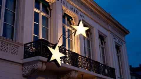 An elegant hotel’s façade, featuring tall windows, floral motifs and an iron balcony, features signs reading ‘HOTEL MERIAN’ and ‘CAFE SPITZ’ and is illuminated by a two illuminated five-pointed stars
