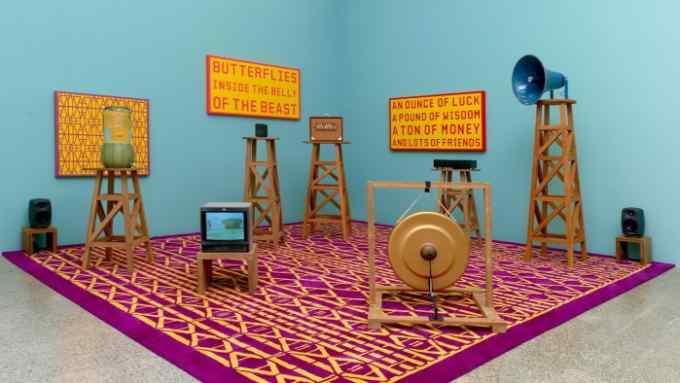 A large yellow and purple patterned carpet against two light blue walls. Large wooden structures like latticed side tables sit on the carpet supporting various objects. Slogan paintings are on the walls.