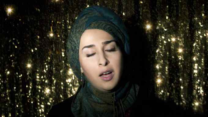 Close-up of a woman in a headscarf singing against a golden curtain