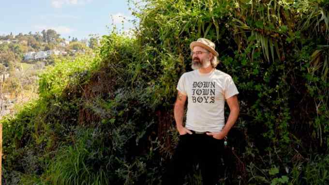 A bearded man with glasses and a tan hat stands in front of a lush, green background of climbing plants and foliage. He is wearing a cream-colored t-shirt with ‘DOWNTOWN BOYS’ printed on it, black pants, and distinctive yellow shoes