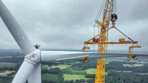 A large  mobile crane lifts a rotor blade to the nacelle of a wind turbine