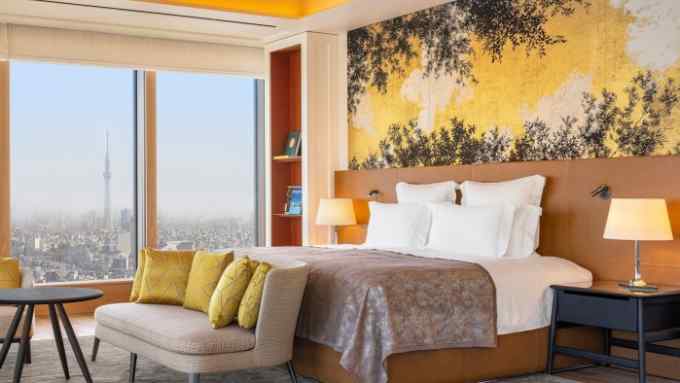 A super-king-size bed, with a gold-throw over it, and pillows stacked behind a headboard in front a wall covered in a yellow and gold botanic mural. The view looks over the city, with the Tokyo Skytree broadcasting and observation tower visible