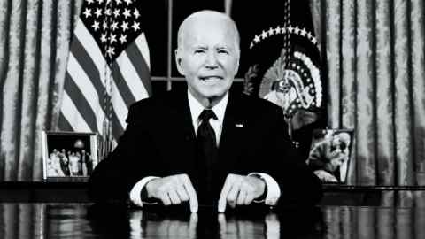 Black and white picture of Biden sitting at his desk in the Oval office with the US flag behind him