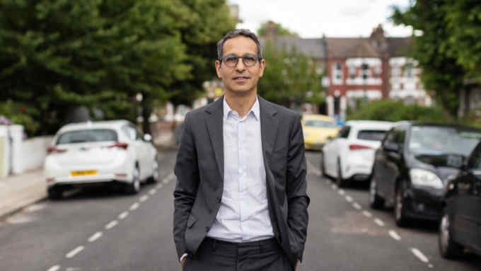 Enver Solomon wearing glasses, blazer and open necked shirt, stands in the road outside his house with his hands in his trouser pockets