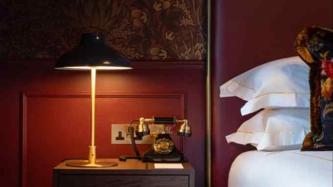 A metal-shaded bedside lamp on a table beside a bed, standing in front of a wood-panelled and brown floral papered wall in a guest room in Sun Street Hotel