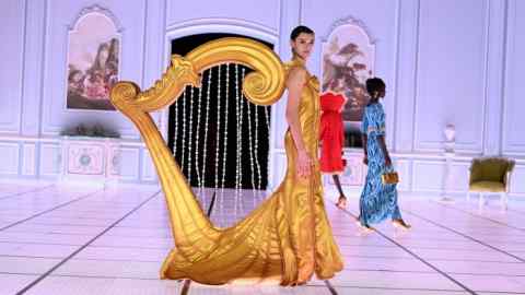 A woman on a catwalk in a golden dress with a sculptural harp-like shape at the back
