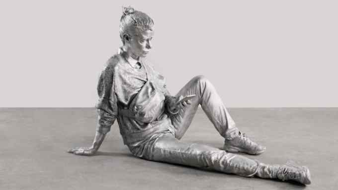 Life-size silver sculpture of a woman in casual clothes relaxing on the ground and looking at her phone