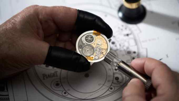 Close-up of a watchmaker’s hands in black gloves assembling a Piaget watch movement, with the intricate parts and schematics visible in the background