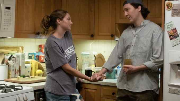 A woman and a man hold hands affectionately in a cluttered kitchen