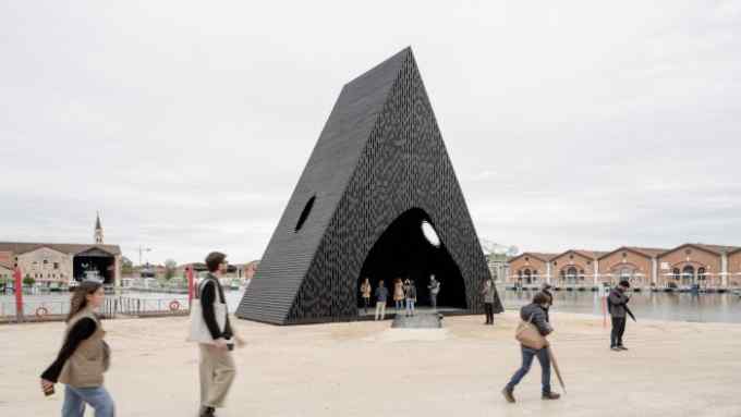 People walk past and cluster under a dark pyramid-shaped building on a waterfront
