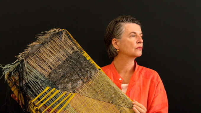 A woman in an orange cotton top, looking severe, holds a panel of yellow and black yarn