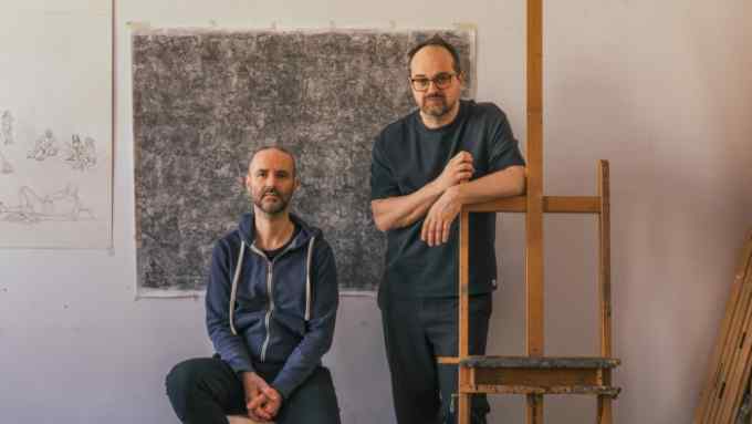 Two middle-aged men, one sitting on a stool on the left, the other leaning on a wooden easel on the right, are captured in their sketch-filled studio